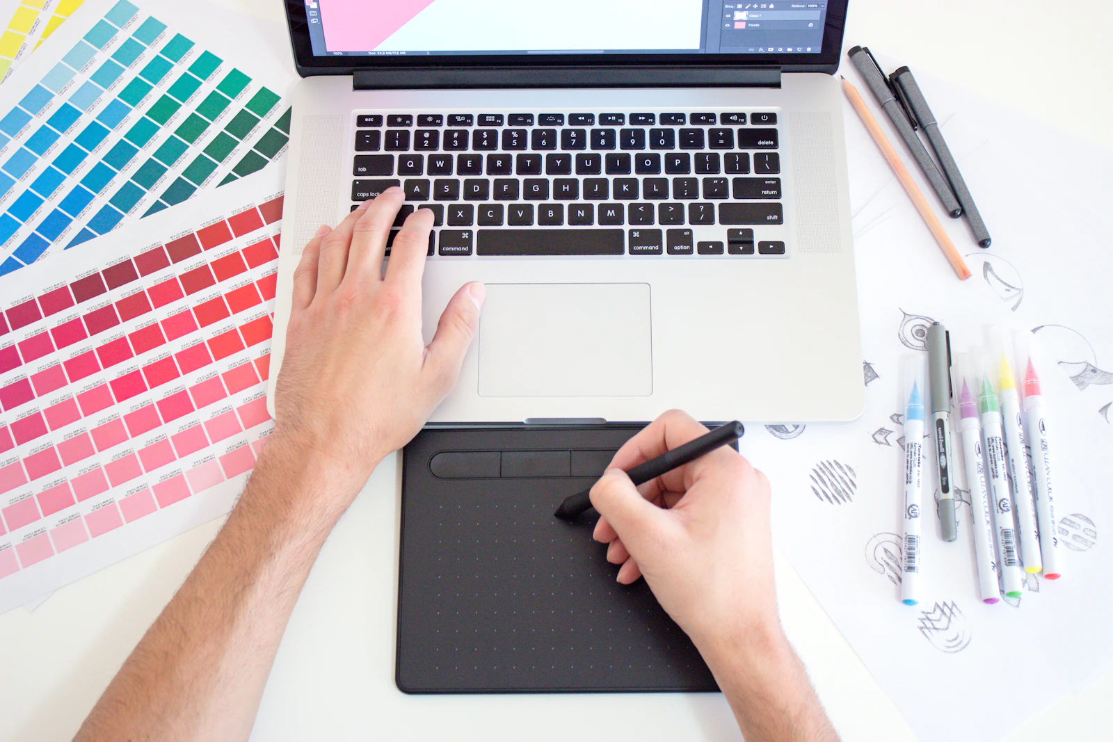 A person using a digital drawing tablet and laptop with color swatches and markers